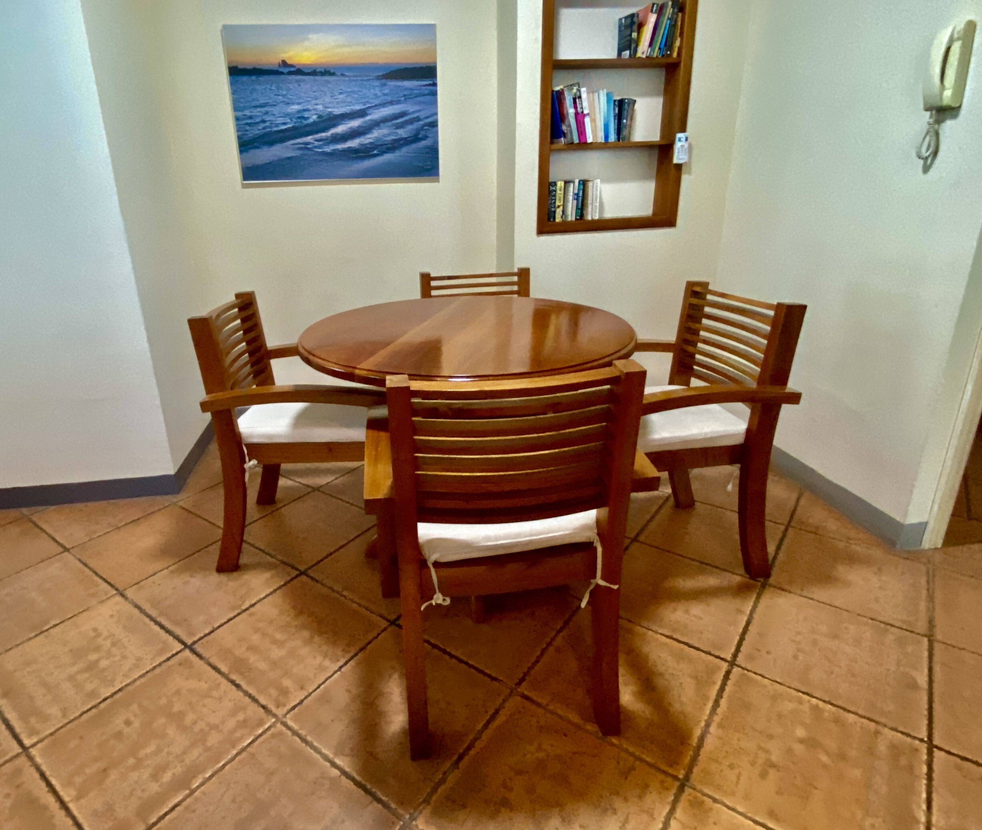 A dining area with a round wooden table and four chairs, a bookshelf with books, and a painting of a sunset over the ocean on the wall.