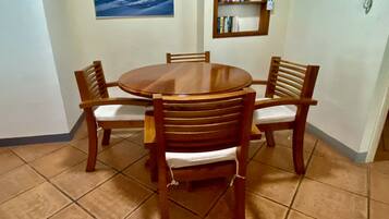 A dining area with a round wooden table and four chairs, a bookshelf with books, and a painting of a sunset over the ocean on the wall.