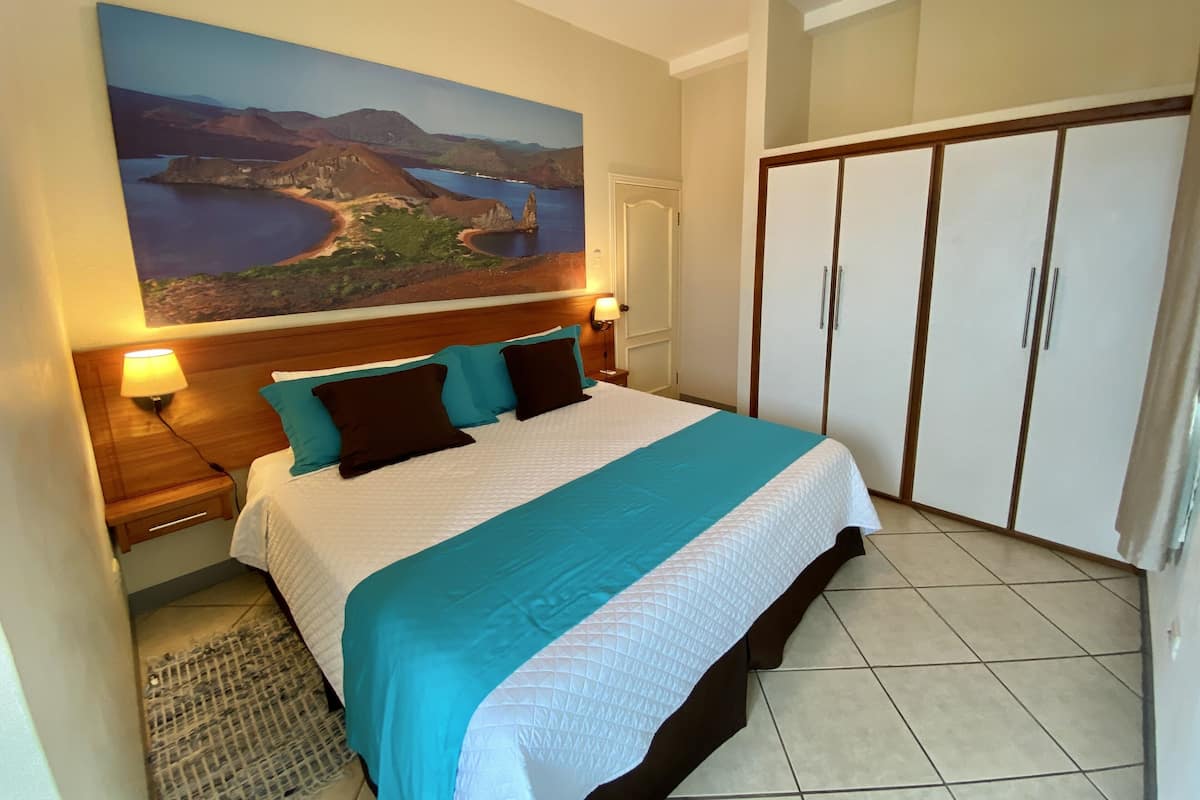 A hotel room with a large bed, a nightstand, a lamp, a wardrobe, and a painting of a landscape on the wall.
