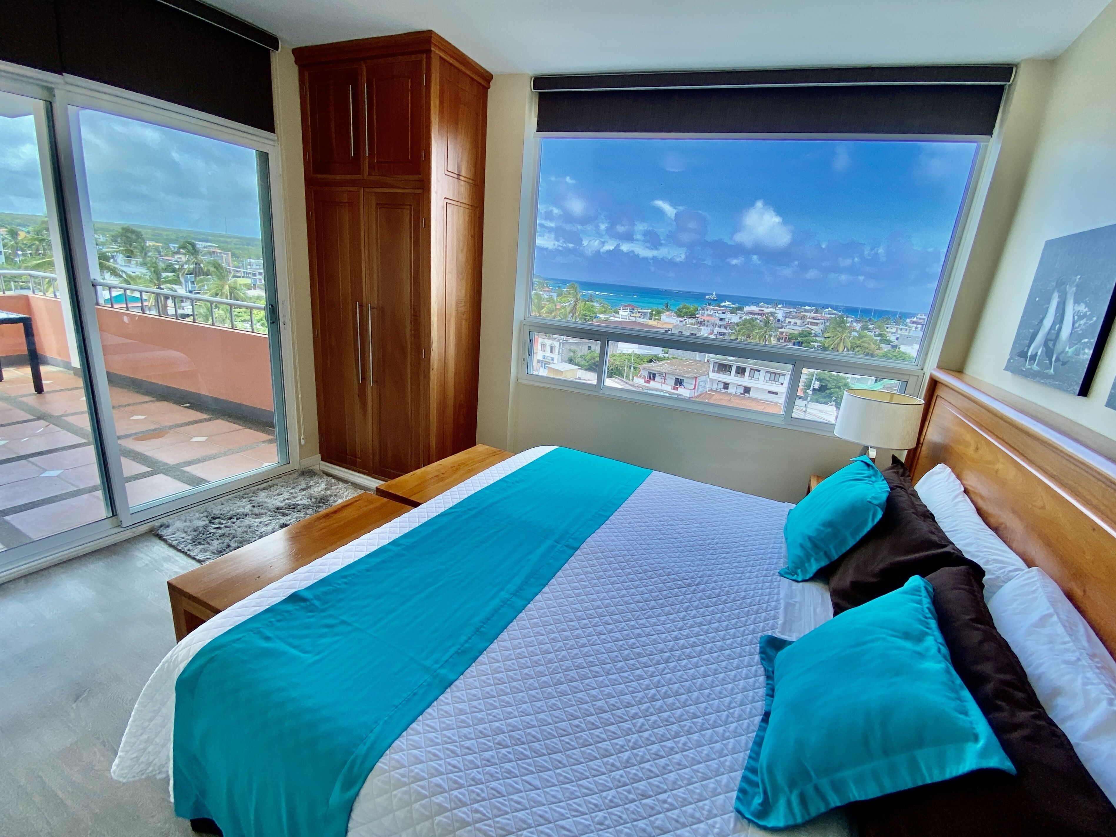 Un dormitorio con una cama grande, un armario de madera, un balcón con vistas a la ciudad y al océano, y una ventana con vistas al océano.