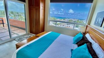 Un dormitorio con una cama grande, un armario de madera, un balcón con vistas a la ciudad y al océano, y una ventana con vistas al océano.