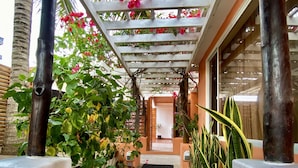A covered walkway with a concrete path, surrounded by plants and flowers, leading to a building entrance.