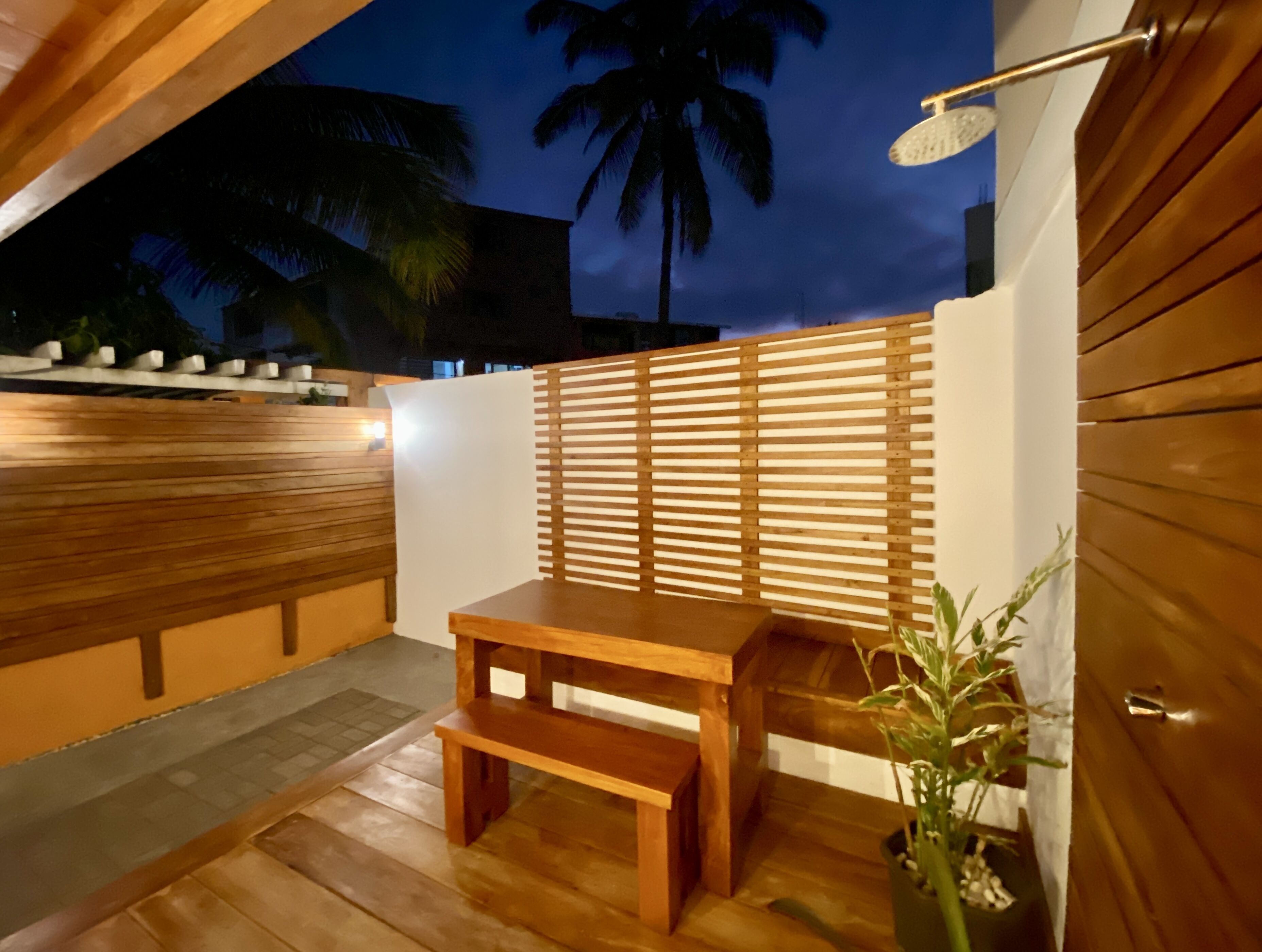 An outdoor seating area with a wooden table and bench, a white wall, and a palm tree in the background.