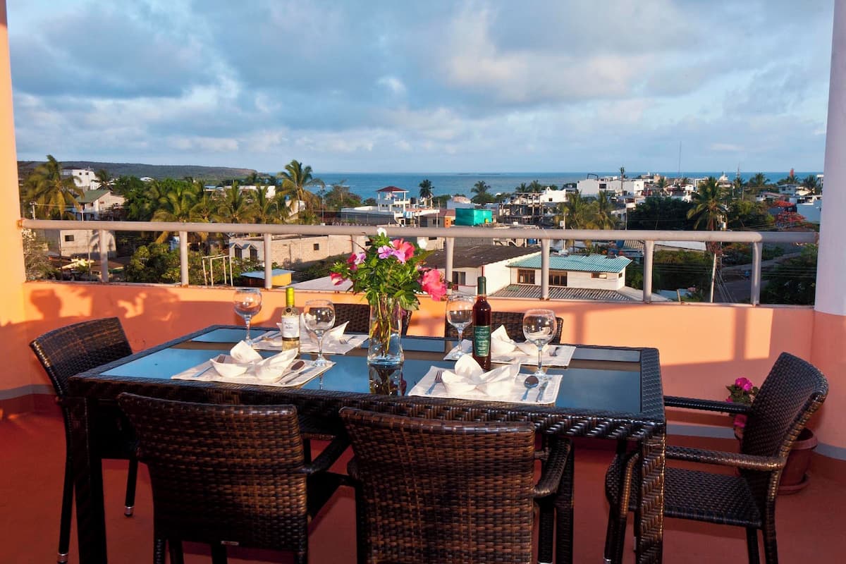 A dining table with chairs, a vase with flowers, and wine glasses set on a balcony overlooking a coastal town.