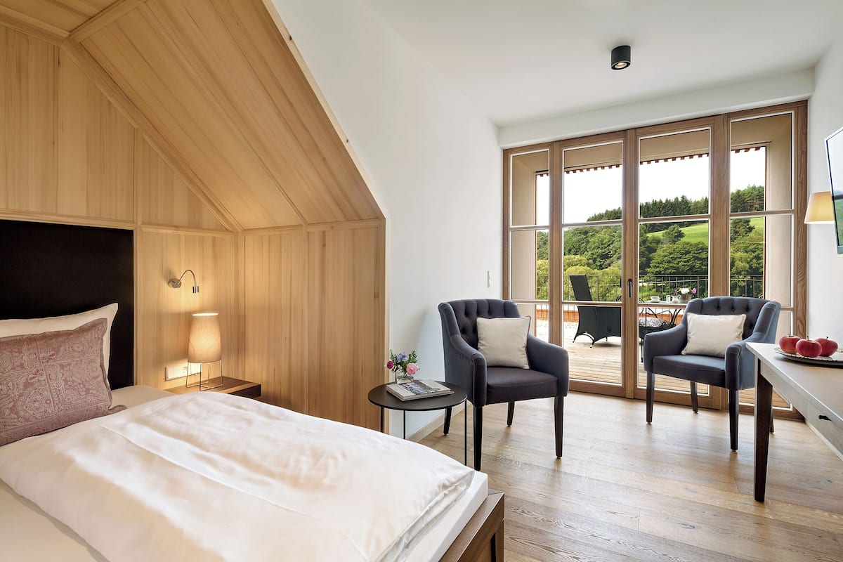 A bedroom with a large bed, two armchairs, a small table with a lamp, and a view of the outdoors through large windows.