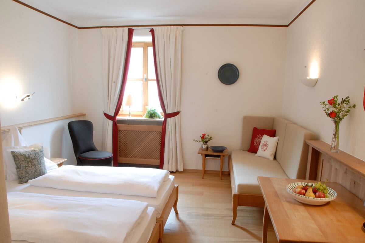 A hotel room with two beds, a sofa, a small table with a bowl of fruit, a chair, a window with curtains, and a wall-mounted light fixture.