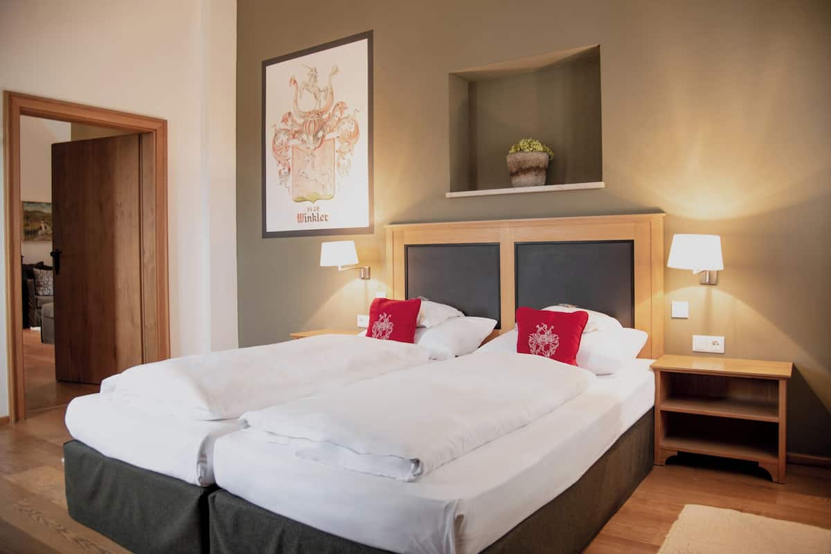A double bed with white linens and red pillows, two bedside tables with lamps, a framed picture on the wall, and a small shelf with a plant.