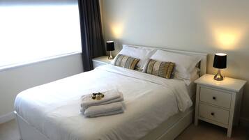 Een opgemaakt bed met witte lakens, twee bedlampjes en een stapel handdoeken op het voeteneinde.