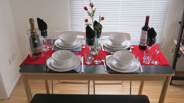 A dining table set with white plates, bowls, and glasses, a bottle of wine, and a vase with red flowers. - City Centre Belfast (Belfast)