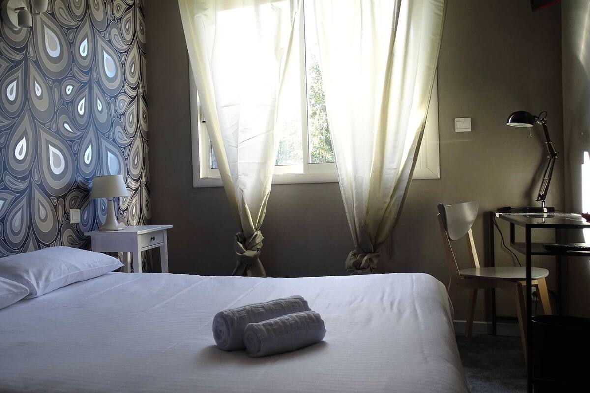 A neatly made bed with a rolled towel, a bedside table with a lamp, a chair, and curtains.