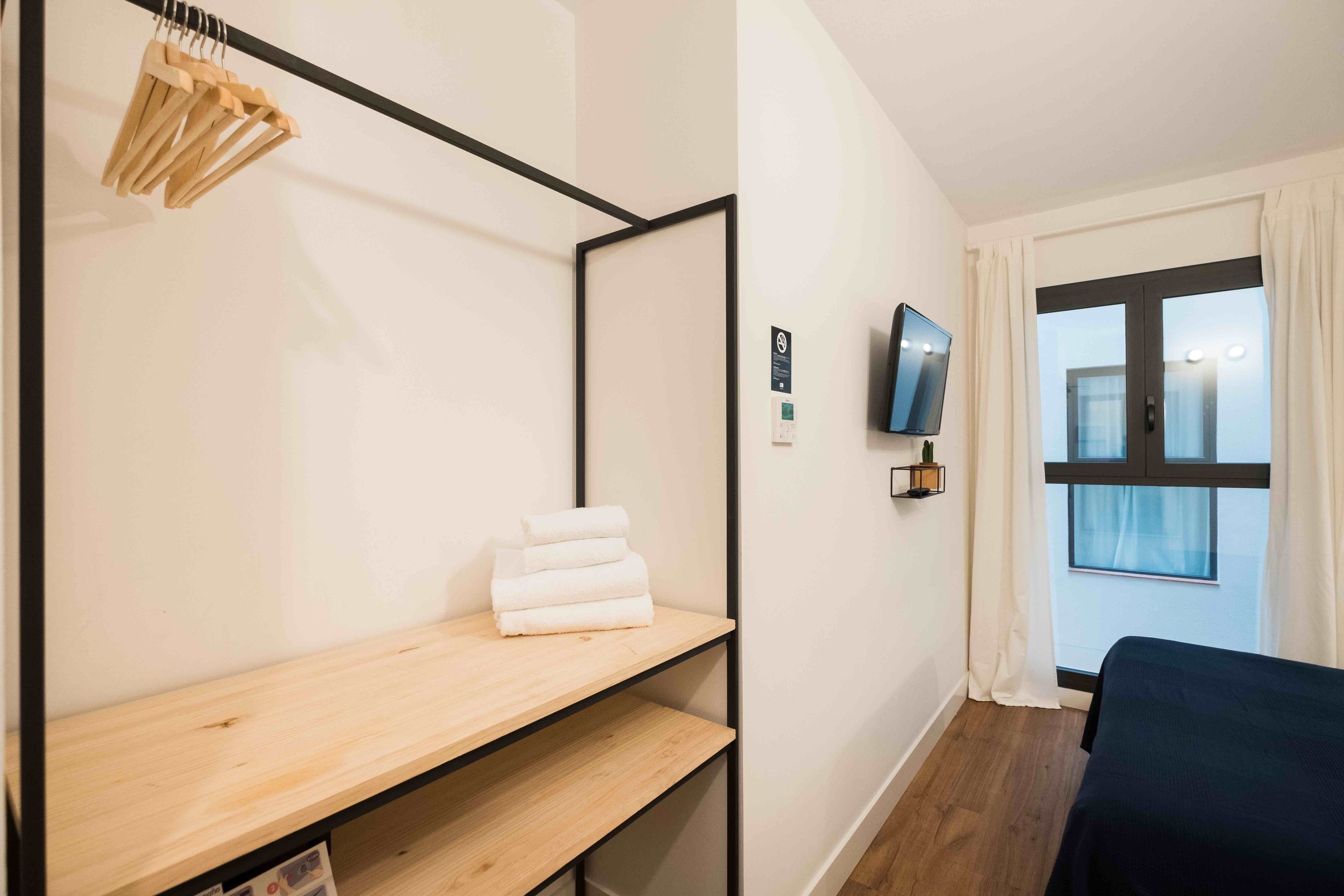 A room with a bed, a TV mounted on the wall, a window with curtains, and a shelf with towels.