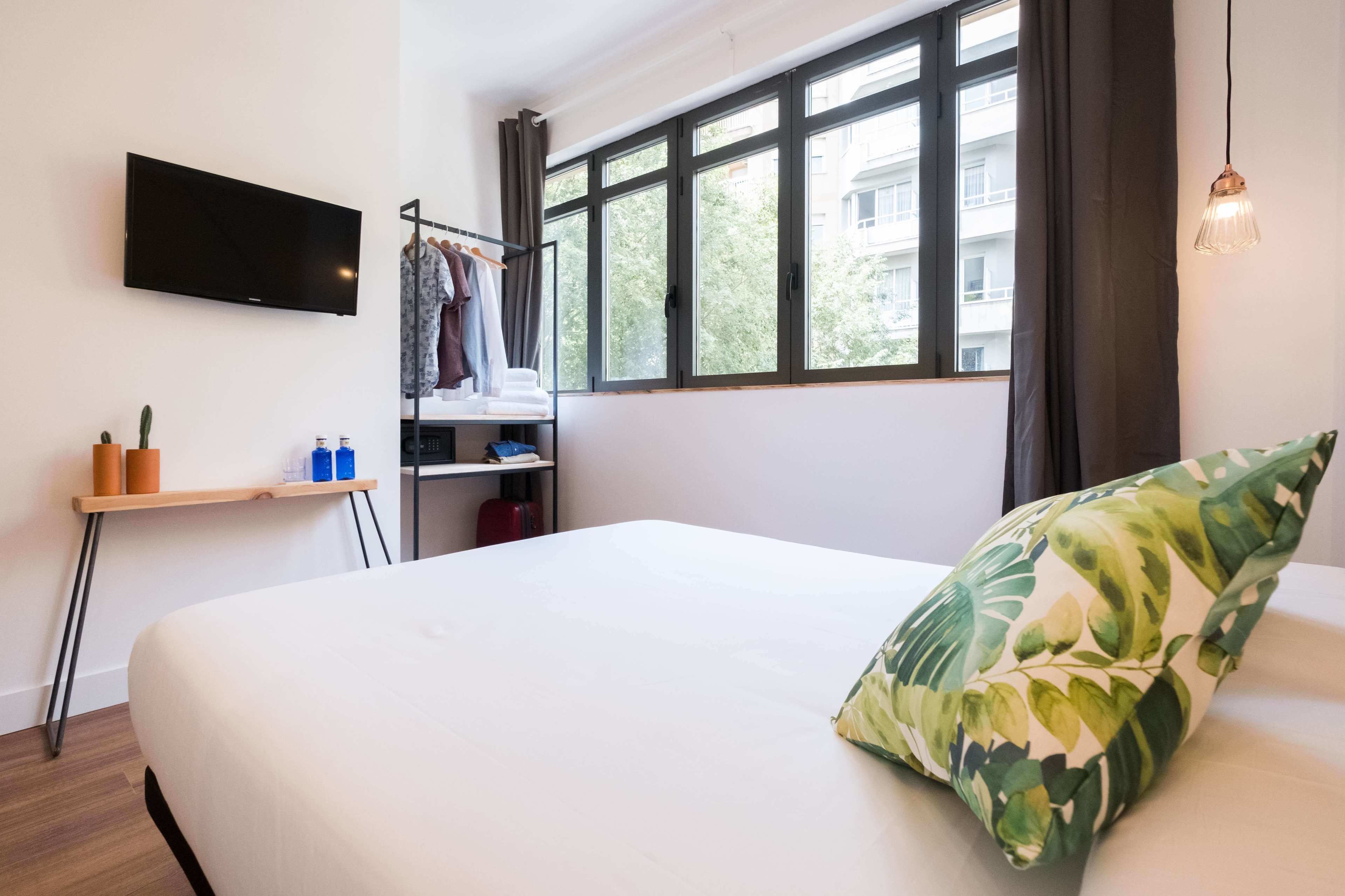 Una habitación de hotel con una cama, un televisor montado en la pared, un ventanal con cortinas, una mesita con un cactus y una lámpara colgante.