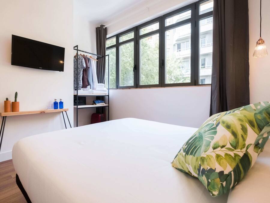 Una habitación de hotel con una cama, un televisor montado en la pared, un ventanal con cortinas, una mesita con un cactus y una lámpara colgante.