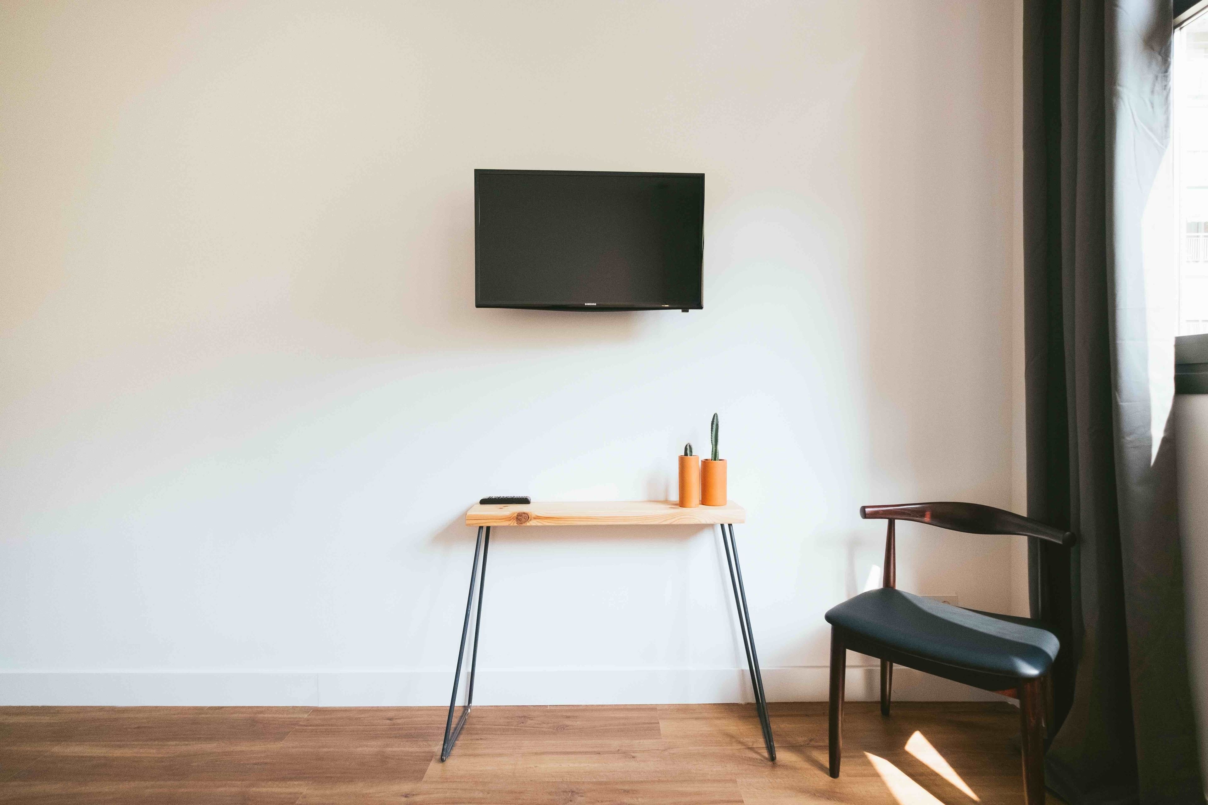 Een kamer met een houten vloer, een aan de muur bevestigde televisie, een klein houten bureau met een stoel en een potplant.