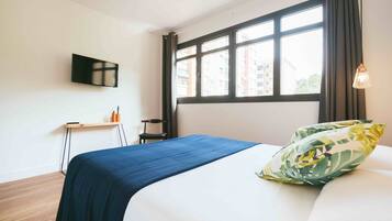 Habitación de hotel con una cama, televisión montada en la pared, ventana con cortinas y una mesita con artículos de aseo.