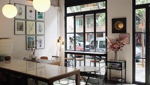Interior de una cafetería con un amplio ventanal, obras de arte enmarcadas y un jarrón con flores.