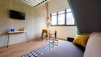 Una habitación moderna con una televisión de pantalla plana en la pared, una mesita con una taza, un sofá con un cojín estampado y una ventana con vistas a edificios.