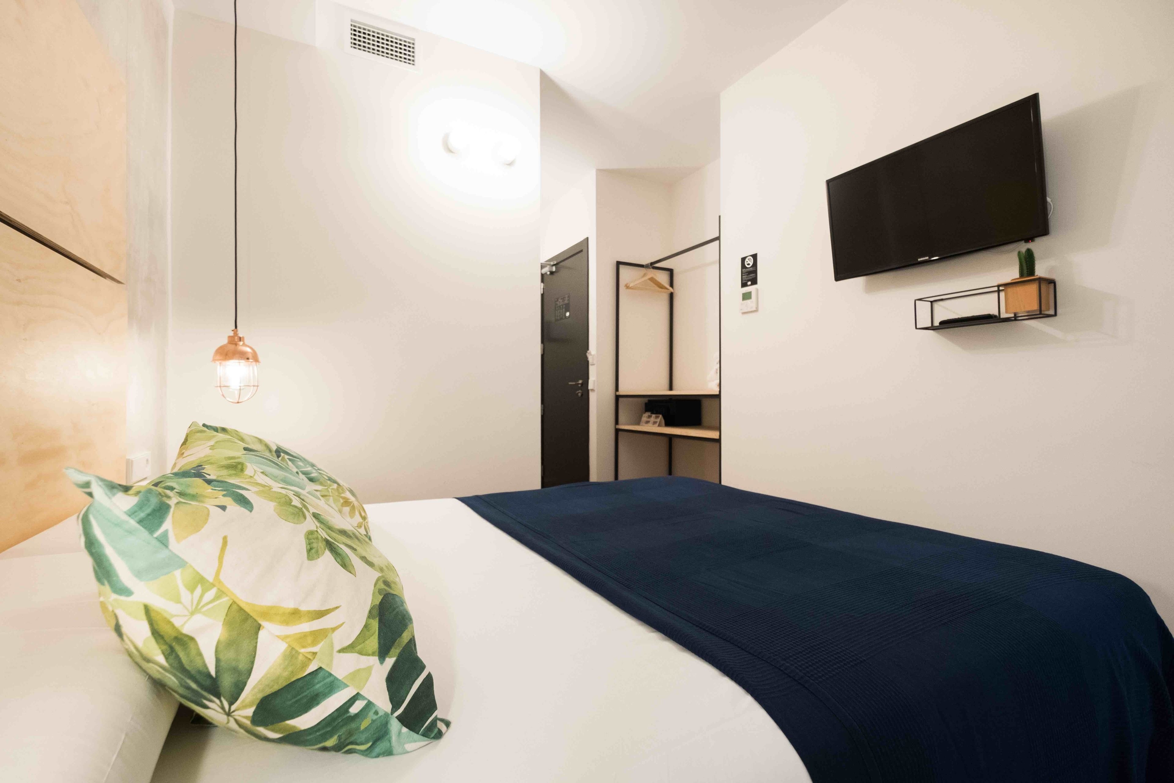 A hotel room with a bed, a hanging light fixture, a mounted television, and a small shelf with a plant.