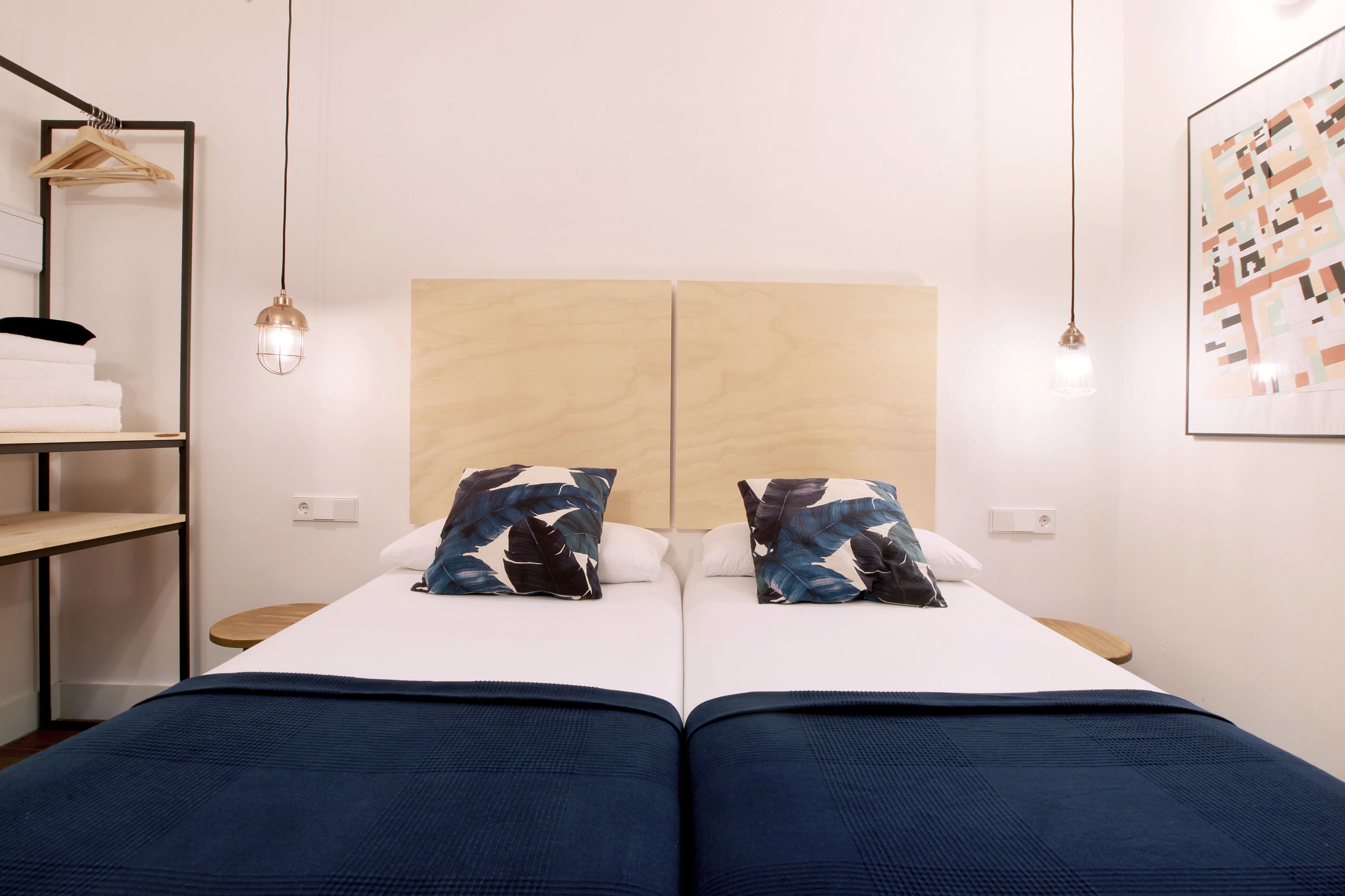 A double bed with a wooden headboard, two pillows, a blue blanket, a hanging light fixture, and a framed artwork on the wall.