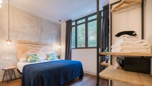 Una habitación de hotel moderna con cabecera de madera, cama bien hecha con cubrecama azul oscuro, mesita de noche con lámpara y ventana con cortinas.