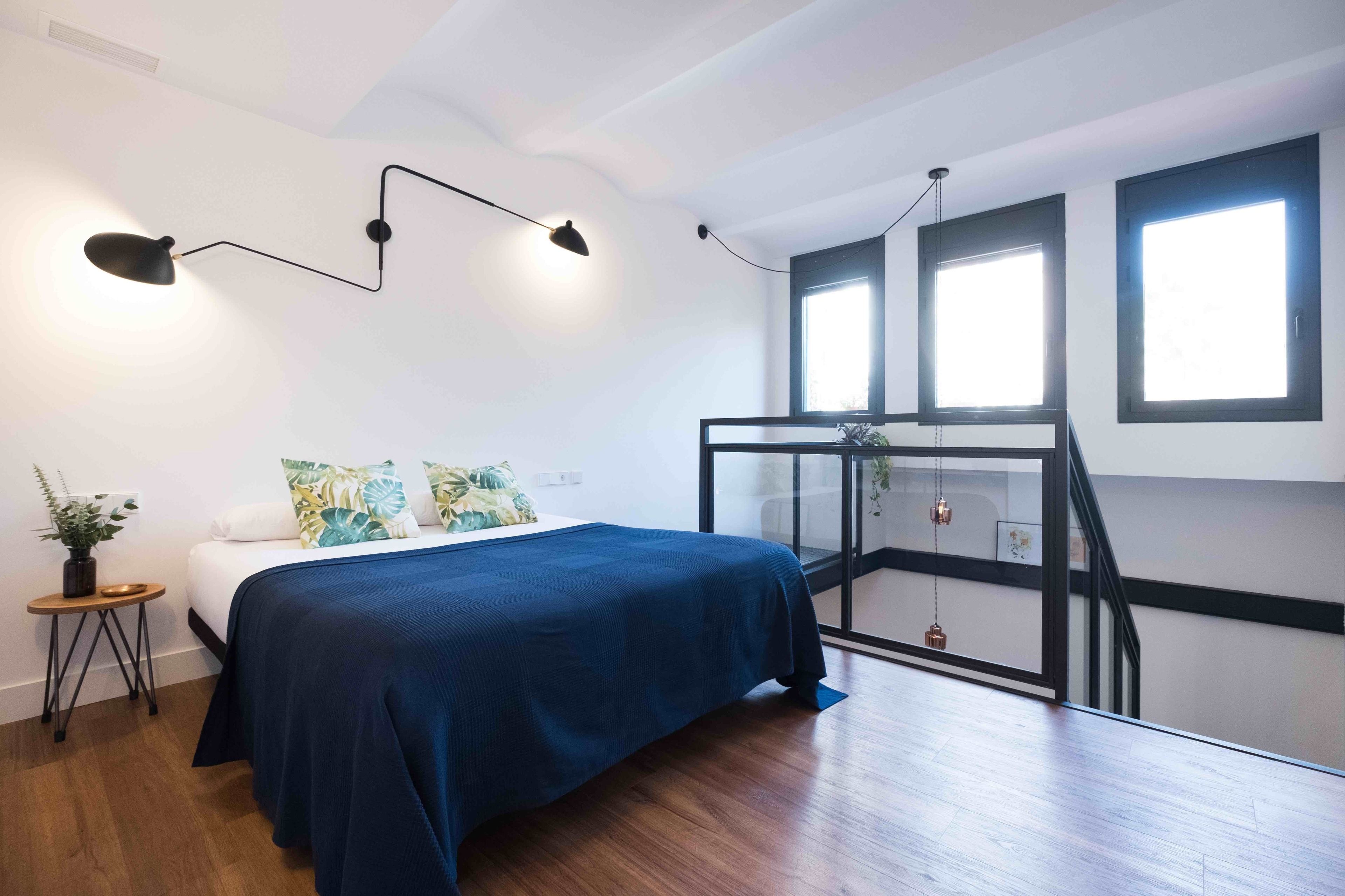 Un dormitorio moderno con una cama, una mesita auxiliar, una planta en maceta y un balcón con barandilla de cristal.