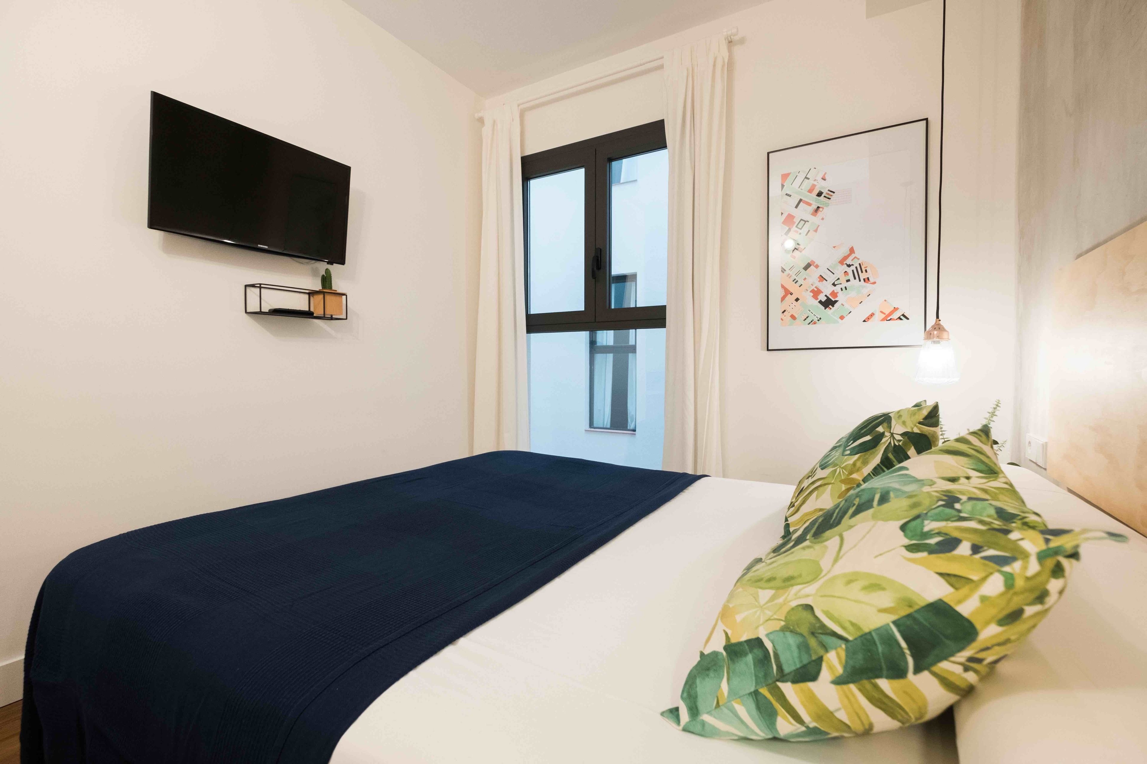 A hotel room with a bed, a flat-screen TV mounted on the wall, a window with curtains, and a framed abstract art piece.