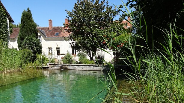 A clear water canal with a stone bridge, surrounded by greenery and a white building with a tiled roof. - Le Jardin de Canaan (Saint-Julien-de-Chedon)