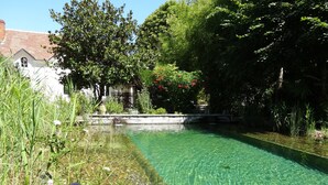 A clear, rectangular pool with a stone border, surrounded by lush greenery and a white building with a red roof in the background. - Le Jardin de Canaan (Saint-Julien-de-Chedon)