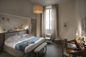 Quarto de hotel com uma cama grande, uma escrivaninha com uma revista, uma cadeira e uma janela com cortinas.