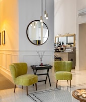 Um lobby de hotel moderno com dois poltronas verdes, um espelho redondo, luminárias pendentes, uma pequena mesa com um vaso e uma kitchenette ao fundo.