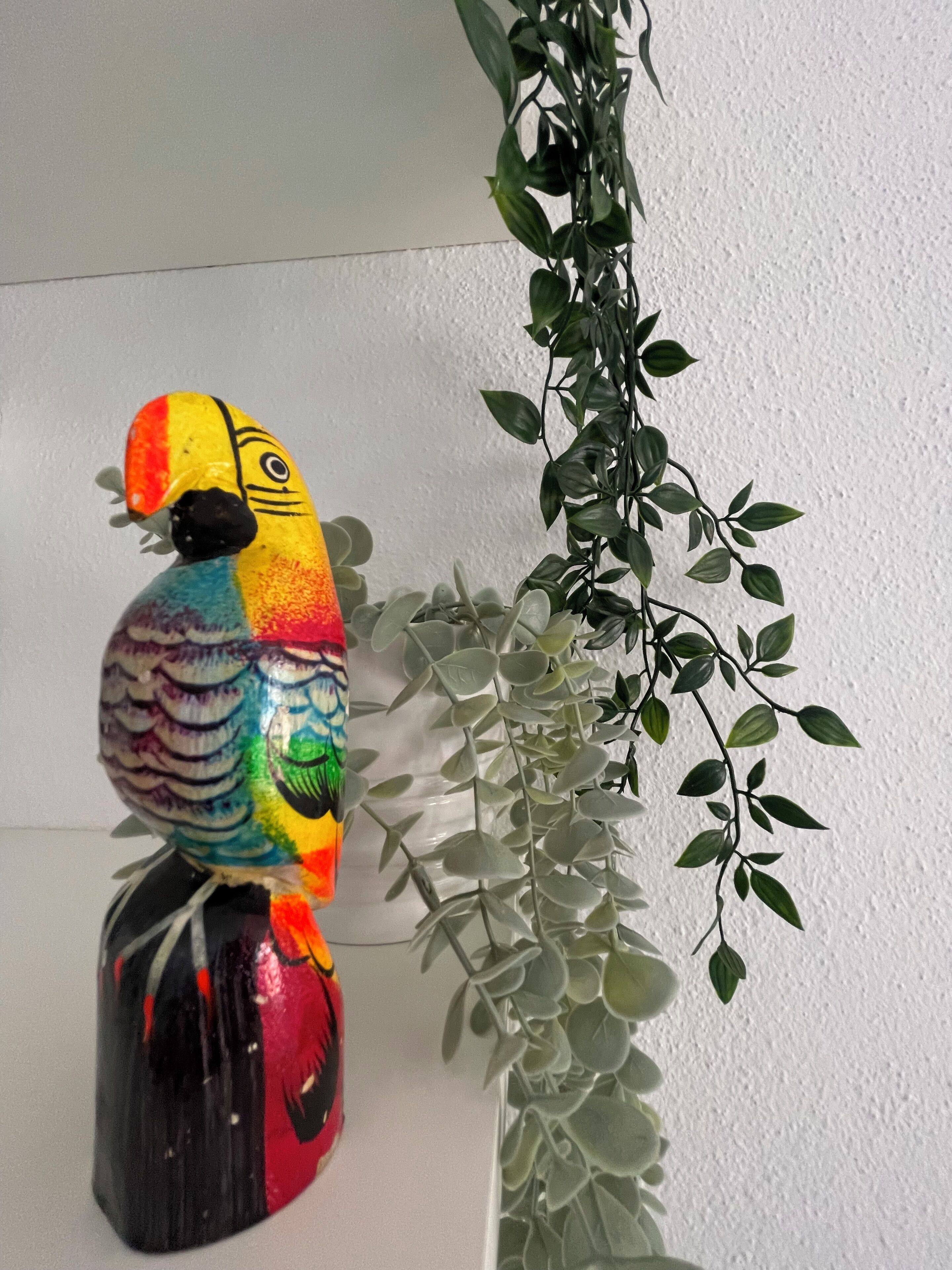 A colorful bird sculpture on a black pedestal next to a white wall and a green plant.
