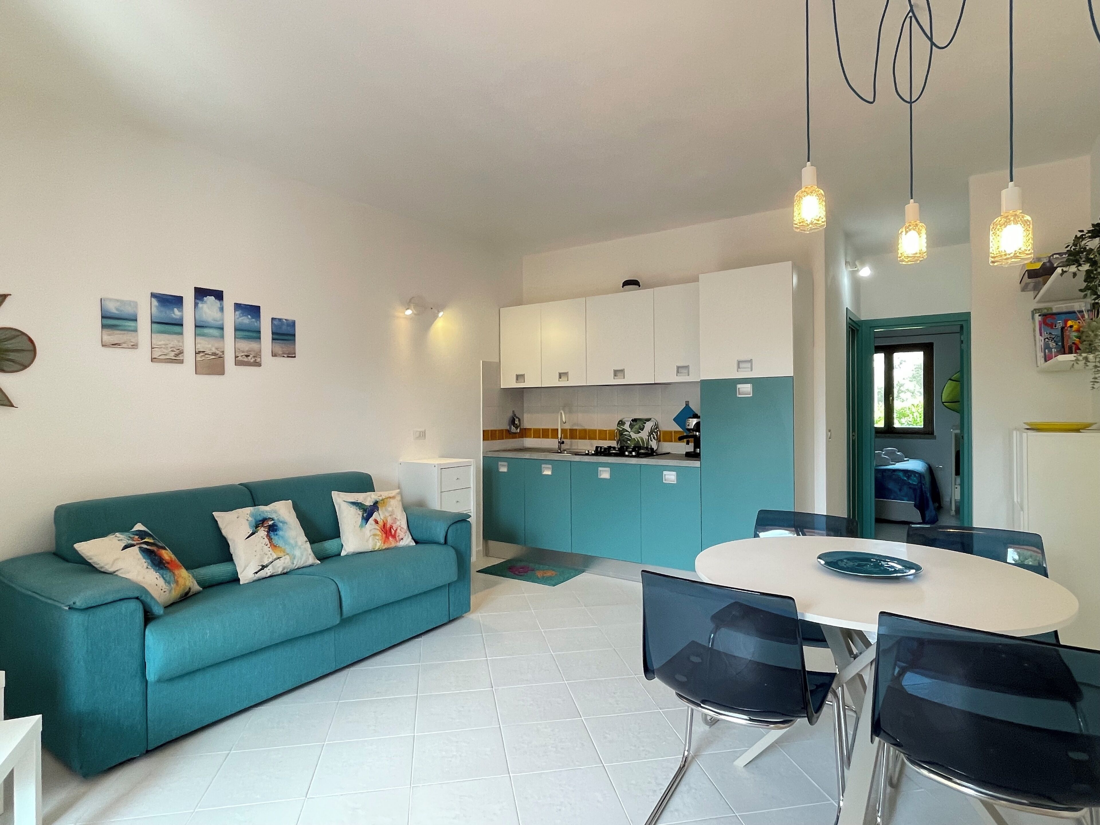 Une cuisine moderne avec des meubles de couleur turquoise, un coin repas avec une table blanche et des chaises noires, et un canapé turquoise agrémenté de coussins colorés.