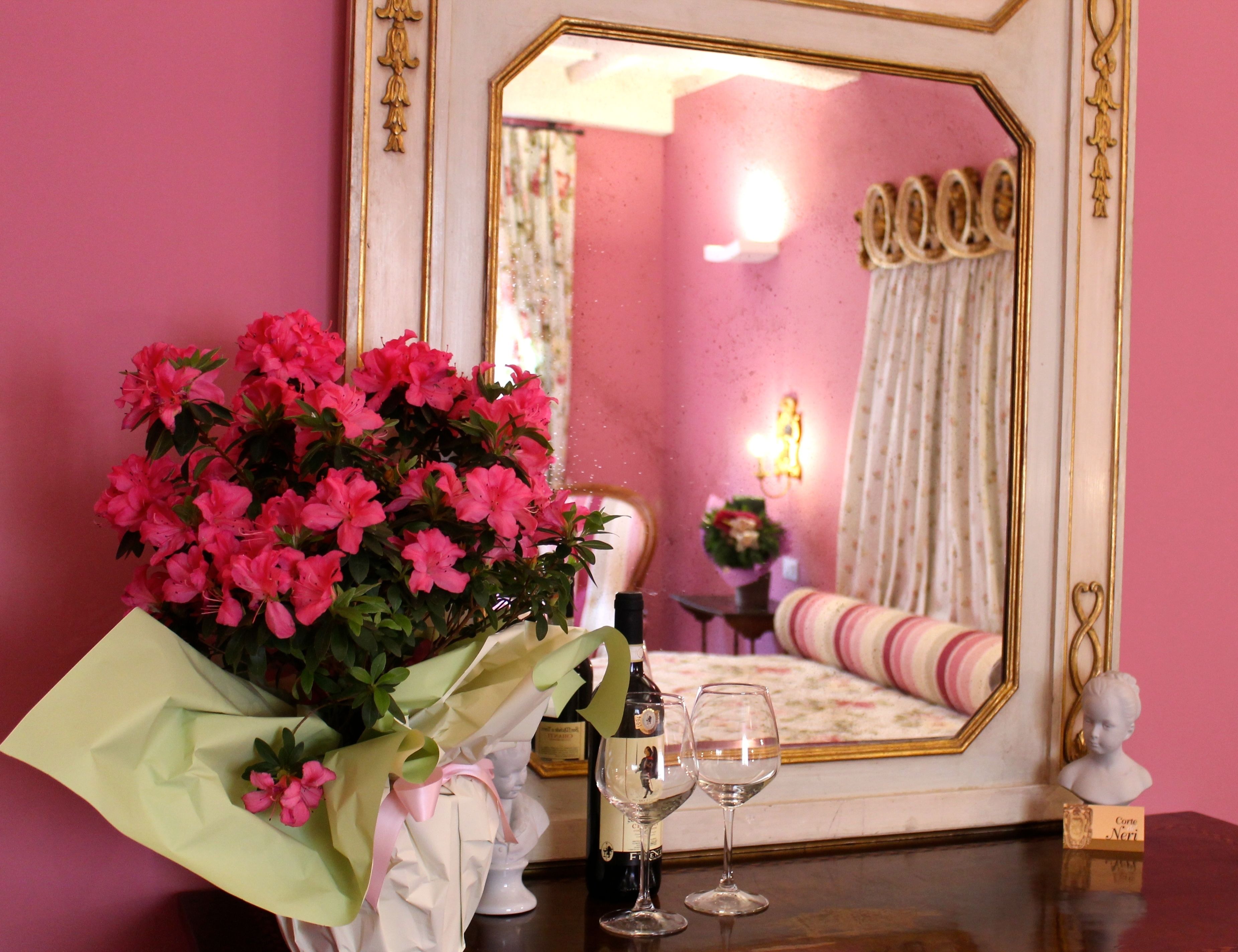 A room with a mirror, a bouquet of pink flowers, two wine glasses, and a bust on a table.