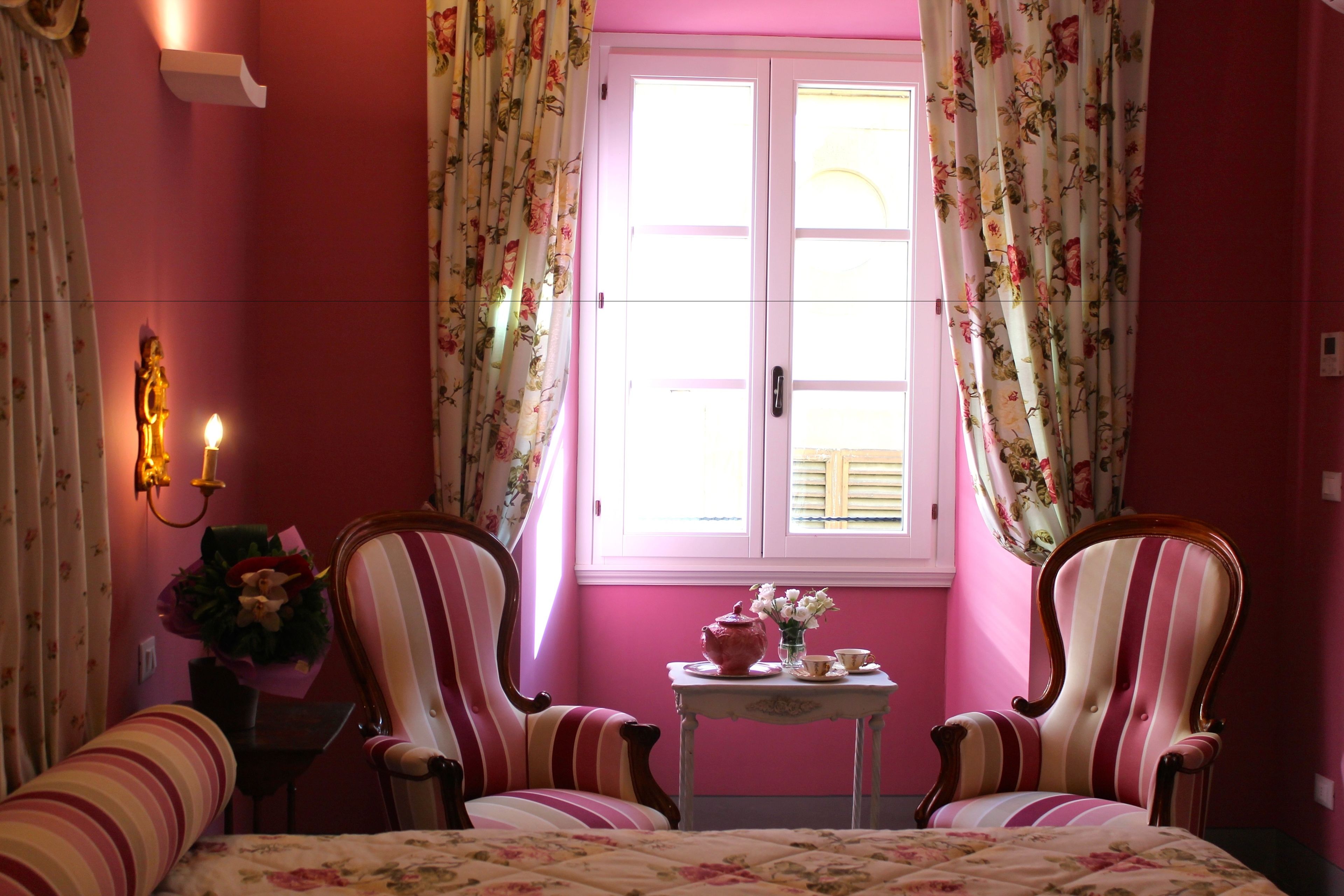A pink-themed room with two striped armchairs, a small table with a teapot and cups, and a window with floral curtains.