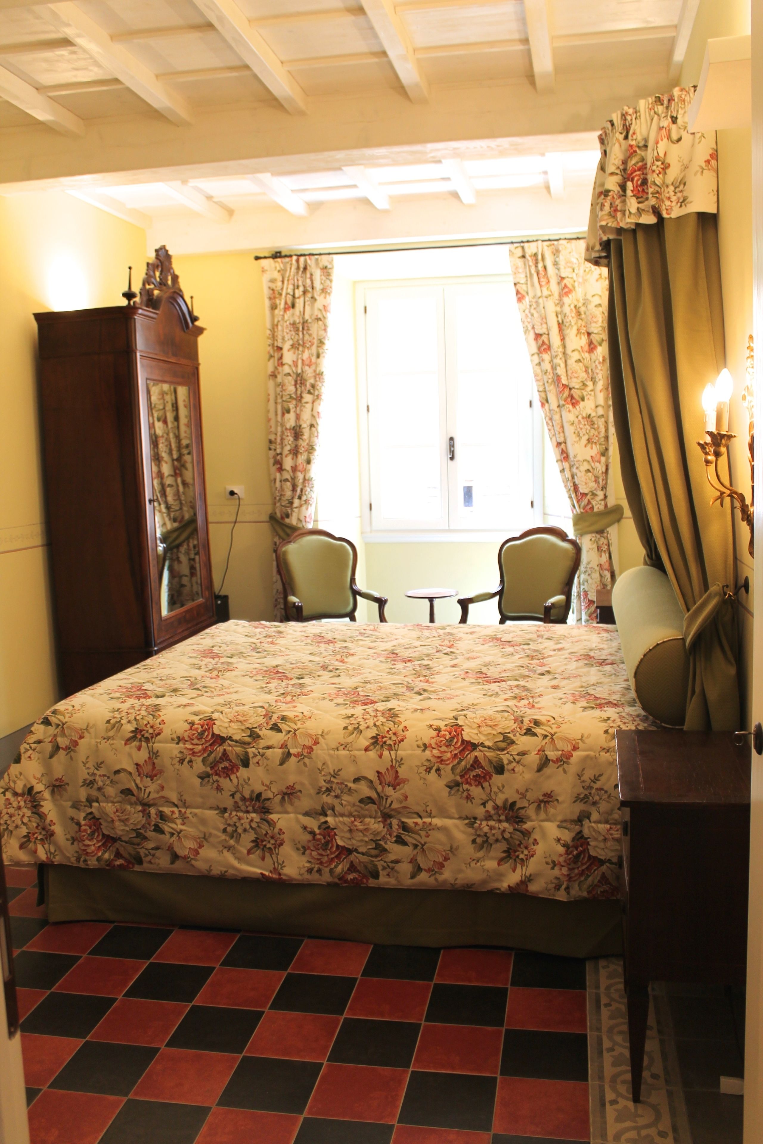 Un dormitorio con colcha estampada, un armario de madera, dos sillas, una mesita y una ventana con cortinas.