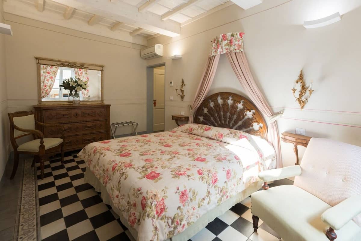 Un dormitorio con suelo de cuadros, colcha estampada, cabecero de madera, espejo, cómoda y una silla.