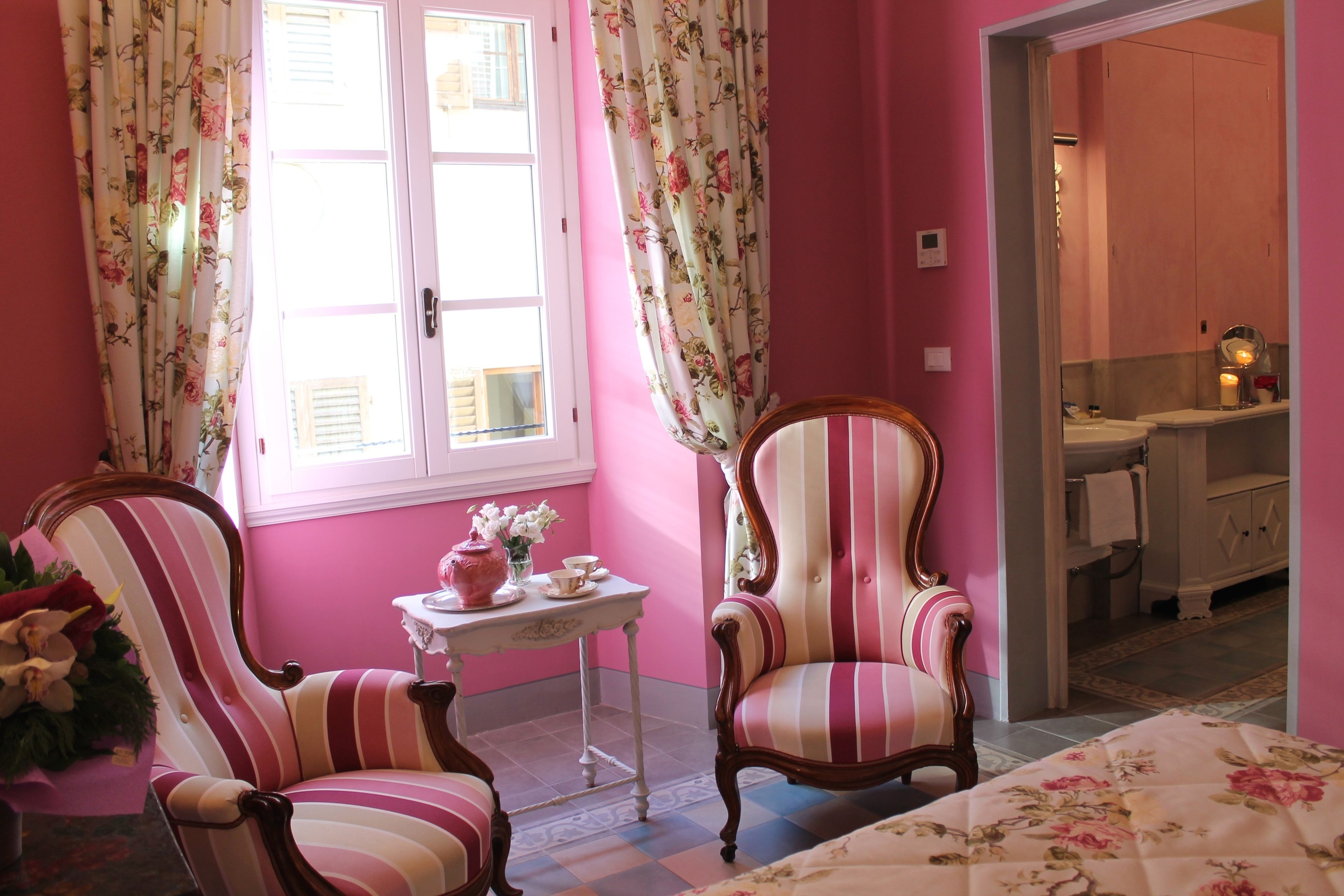 Una habitación con paredes rosas, dos sillones a rayas, una mesita con un jarrón de flores y vistas a un baño con lavamanos y espejo.