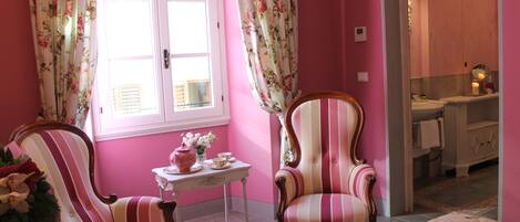 Una habitación con paredes rosas, dos sillones a rayas, una mesita con un jarrón de flores y vistas a un baño con lavamanos y espejo.