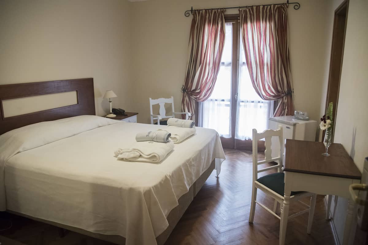 Habitación de hotel con una cama grande, un escritorio con silla, una mesita de noche y un ventanal con cortinas.
