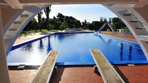 A swimming pool with a slide, surrounded by a covered area with benches and a view of trees and a grassy area.