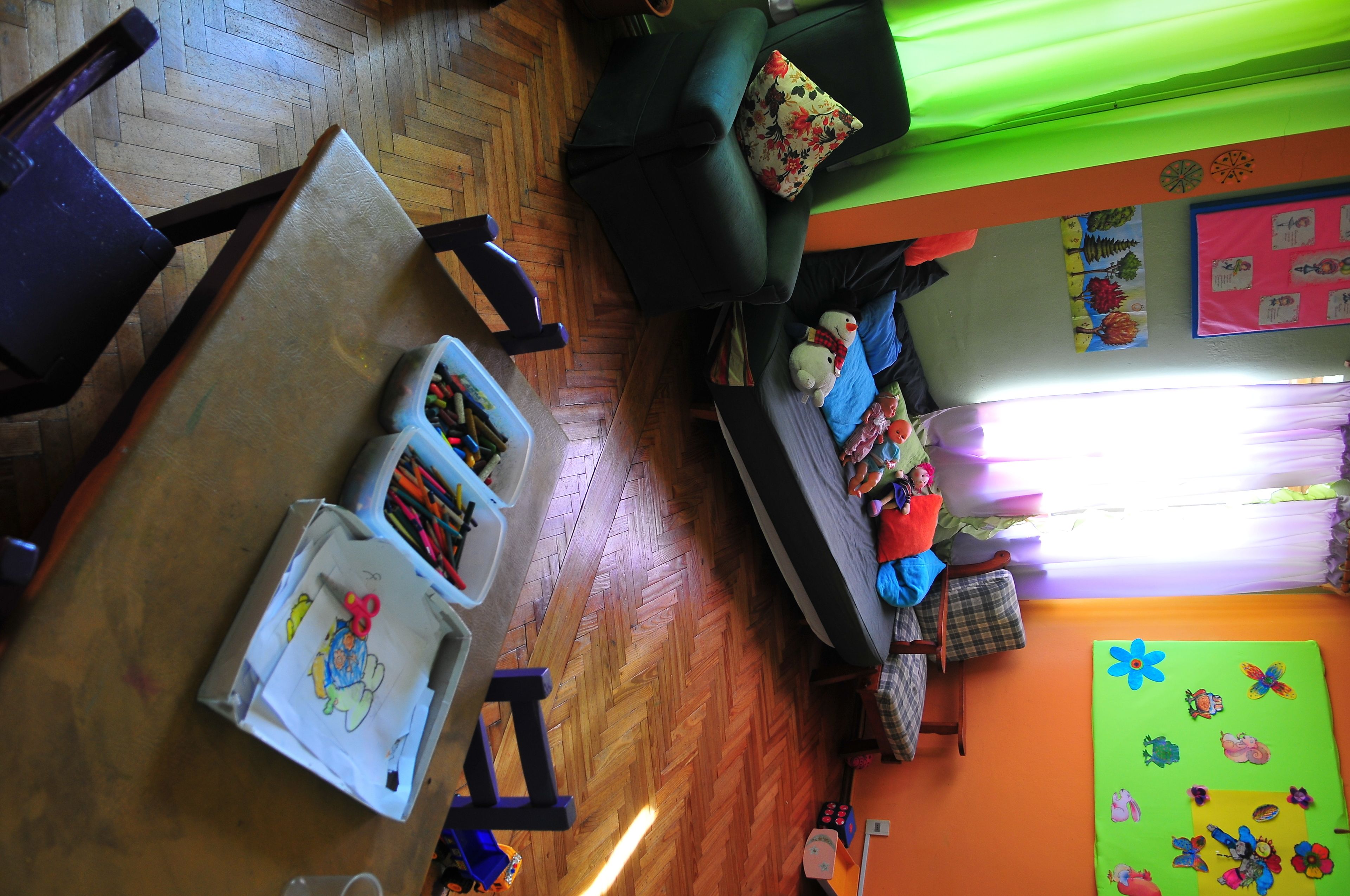 A room with a wooden floor, a green sofa with floral cushions, a table with a white container and a pair of scissors, and a colorful wall decoration.