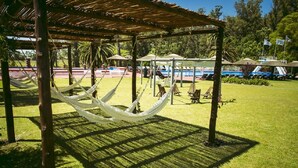A hammock under a wooden pergola in a grassy area with lounge chairs and umbrellas.