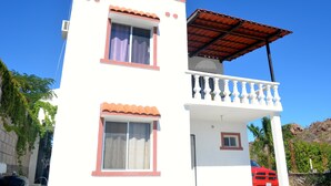 A two-story building with a balcony, a car parked in front, and a mountain in the background. - Las Glorias LG104 1 Bedroom Apartment By Seaside San Carlos (San Carlos)