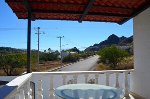 Uma varanda com guarda-corrimão branco, mesa de vidro e vista para uma estrada que serpenteia até as montanhas.