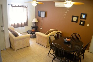 Uma sala de estar com dois sofás, uma mesa de jantar com cadeiras, uma televisão fixada na parede e ventiladores de teto.