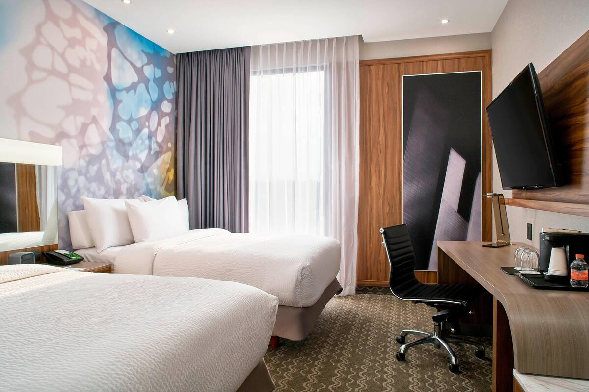 Une chambre d’hôtel avec deux lits, un bureau avec un ordinateur, un téléviseur à écran plat et une grande fenêtre avec des rideaux.