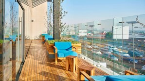 Un espace de détente extérieur moderne, doté d’un plancher en bois, de sièges bleus et de murs en verre offrant une vue sur une autoroute très fréquentée.