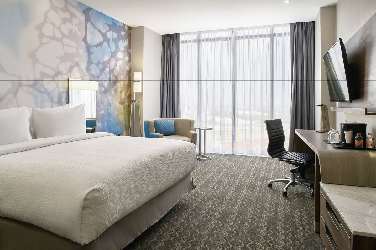 Ein Hotelzimmer mit einem großen Bett, einem Schreibtisch, einem Sessel, einem Fernseher und Blick auf die Stadt durch große Fenster.
