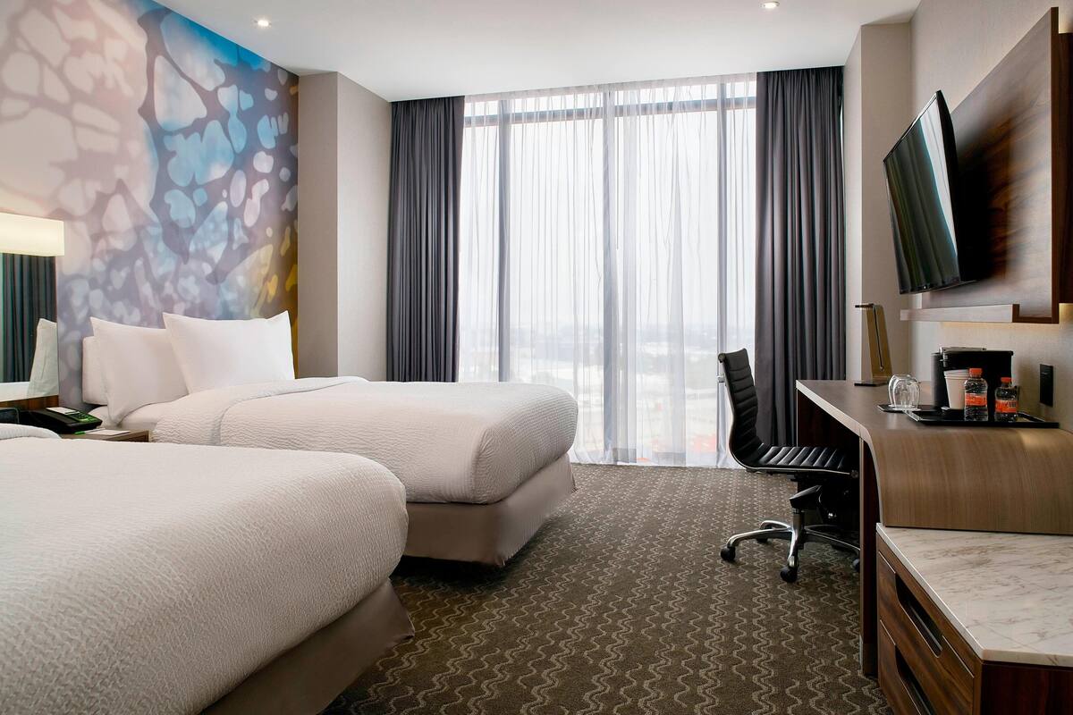 Une chambre d’hôtel avec deux lits, un bureau, une chaise, une télévision et une grande fenêtre avec des rideaux.