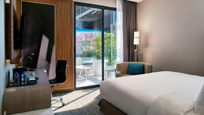 Une chambre d’hôtel avec un grand lit, un bureau avec un ordinateur, une chaise et une vue sur le paysage urbain, visible à travers une porte vitrée.
